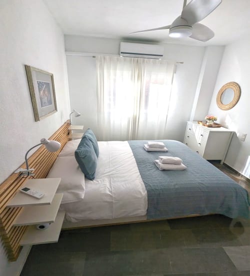 Dormitorio luminoso con cama doble, ropa de cama azul, ventilador de techo y decoración minimalista en apartamento