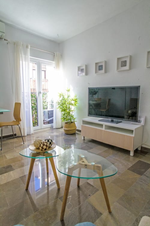 Sala de estar luminosa con televisor, mesas de vidrio, planta decorativa y salida a balcón en apartamento turístico.
