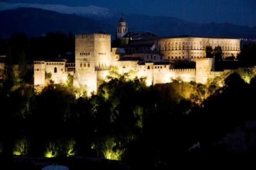 Vista nocturna de la Alhambra iluminada, con la Sierra Nevada de fondo, destacando la majestuosidad del monumento.