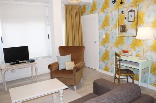 Salón con sofá marrón, sillón a juego, pared de flores, mesa de centro blanca y TV. Cortinas doradas y decoración moderna.