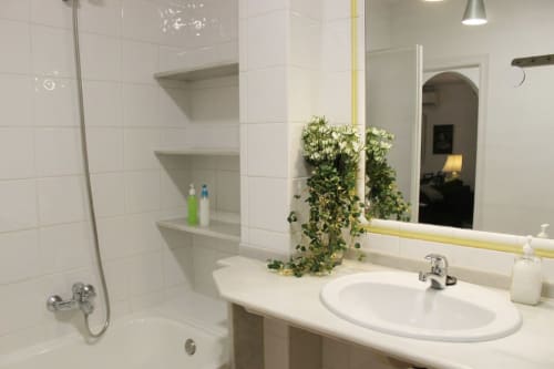 Baño moderno con bañera, ducha, lavamanos, espejo y planta en apartamento de Granada. Azulejos blancos y diseño minimalista.