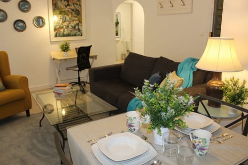Salón moderno con sofá marrón, sillón amarillo, mesa de cristal y comedor. Plantas, cuadros y ambiente acogedor en un apartamento en Granada.