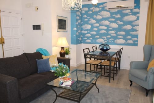 Salón moderno con sofá, sillones, mesa de centro y comedor. Pared con papel pintado de nubes y cortinas doradas. Diseño luminoso y acogedor.