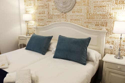 Habitación con cama doble, ropa de cama blanca, cabecero de madera y pared con nombres de ciudades. Mesitas de noche con lámparas y toallas limpias.

