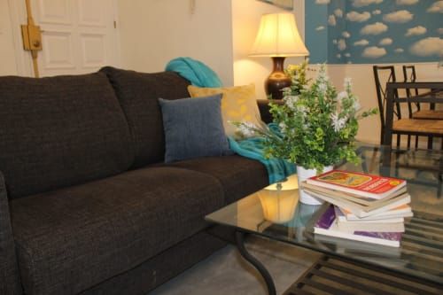 Salón con sofá marrón, cojines, mesa de centro de cristal con libros y decoración floral. Pared con papel pintado de nubes. Ambiente acogedor.
