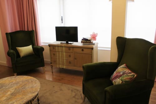 Rincón acogedor con sillones verdes, cortinas rosas, cómoda clásica, televisor y detalles decorativos elegantes.