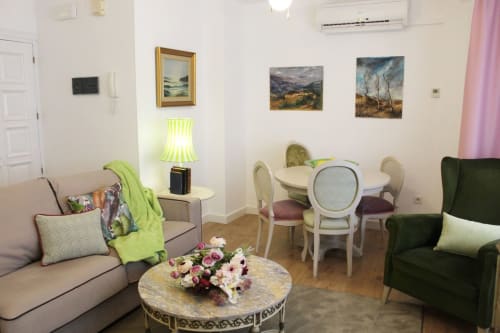 Salón con sofá beige, sillón verde, mesa redonda con sillas clásicas, cuadros coloridos y decoración floral elegante.