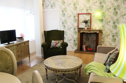 Salón elegante con papel floral, chimenea decorativa, sillón verde, y detalles vintage en tonos cálidos y naturales.