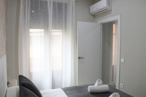 Habitación luminosa con cortinas blancas, aire acondicionado, puerta abierta y detalles modernos en tonos neutros.