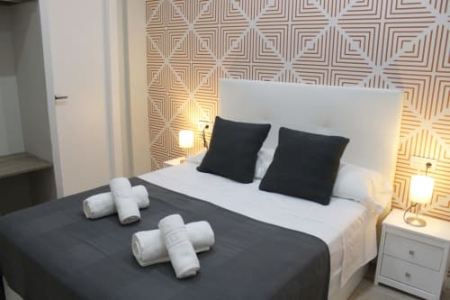 Habitación moderna con cama doble, cojines oscuros, papel tapiz geométrico y luces cálidas en mesitas blancas.
