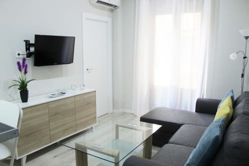 Salón moderno con sofá gris, mesa de cristal, mueble de madera clara, televisor y decoración minimalista.