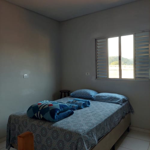 Quarto claro da Pousada Serra Azul em Juquiá com cama, roupa de cama azul e janela com vista para a cidade