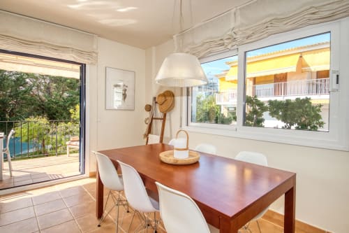 Modern dining room with wooden table, white chairs, and pendant lamp overlooking garden view in Calella apartment