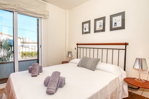 Modern bedroom with wood and metal bed, white linens, purple towels, framed artwork, and balcony view in Calella apartment