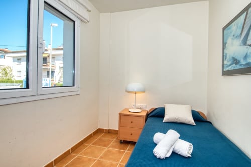 Cozy bedroom in Calella apartment with blue bed, wooden nightstand, lamp, and window overlooking garden.