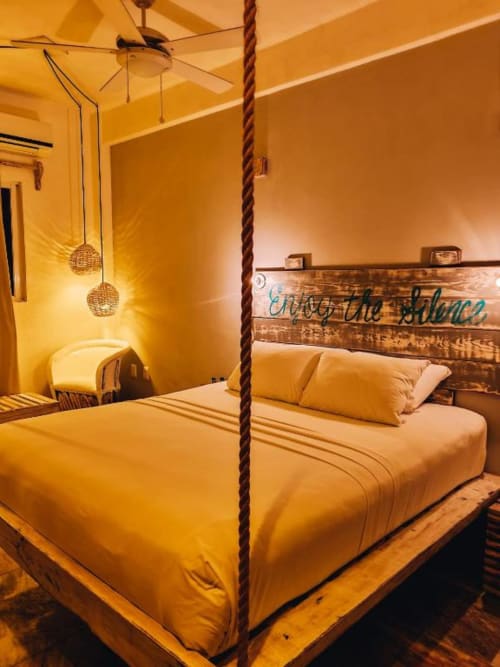 Habitación de tema amarillo en el Hotel 40 Cañones de Mahahual con cama suspendida por cuerdas, iluminación colgante y cabecero de madera