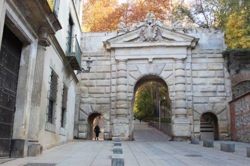 "Puerta de las Granadas" de piedra con arco central y dos laterales, decorada con un escudo, en un entorno arbolado