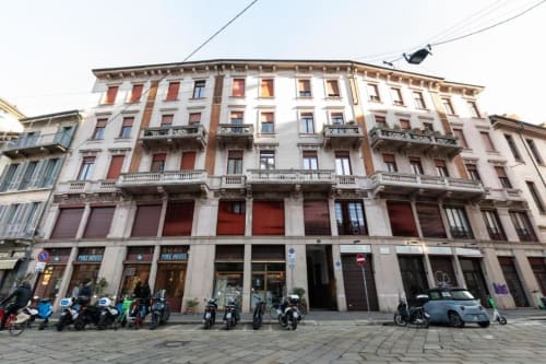 Historische Fassade der Magnolia Apartments Mailand mit roten Türen, Erdgeschossläden und geparkten Rollern in der Stadt