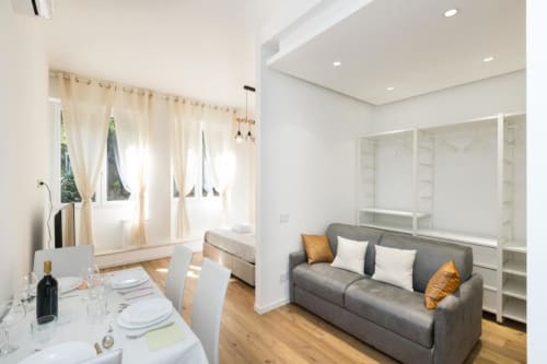 Современная студия в Magnolia Apartments Milan: серый диван, обеденная зона, кровать и встроенные полки в светлом современном интерьере