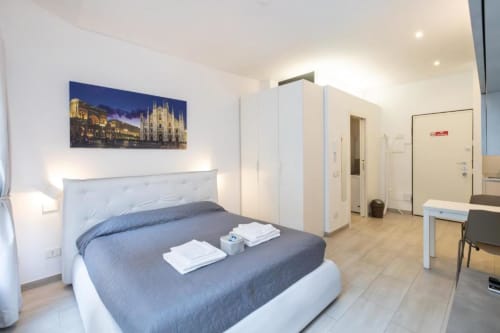 Modernes Schlafzimmer im Magnolia Apartments Milan mit blauer Bettwäsche, weißem Kopfteil, Stadtblick und Küchenzeile