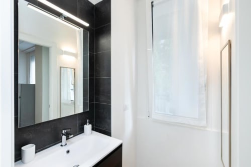 Modernes Badezimmer in den Magnolia Apartments Mailand mit weißem Waschbecken, schwarzer Fliesenwand, beleuchtetem Spiegel und Glasduschkabine