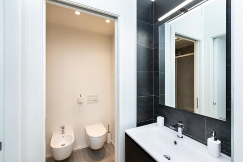 Modernes Badezimmer in Magnolia Apartments Milan mit schwarzen Kacheln, weißem Waschbecken, beleuchtetem Spiegel und separatem Bereich