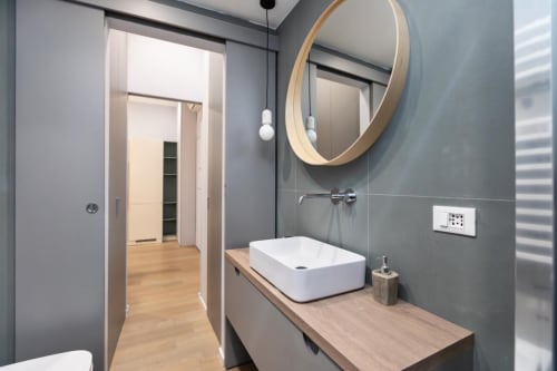 Современная ванная комната в Magnolia Apartments Milan с серой керамической плиткой, деревянной тумбой, белой раковиной и круглым зеркалом
