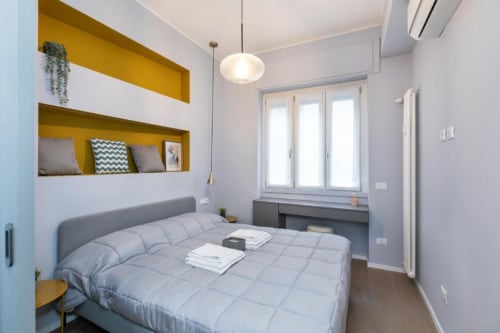 Modernes Schlafzimmer in den Magnolia Apartments Mailand mit grauem Bett, Senf-Regalen, Pendelleuchte und Stadtblick