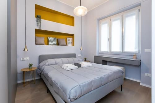 Modernes Schlafzimmer in der Magnolia Apartments Mailand mit grauem Bett, gelben Regalen, Pendelleuchten und Stadtblick
