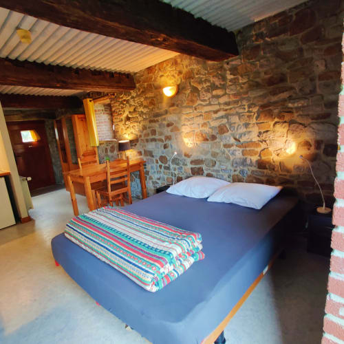 Rustieke stenen slaapkamer in La Ferme De Werpin met zichtbare balken, blauw bed, gekleurde deken en tuinuitzicht