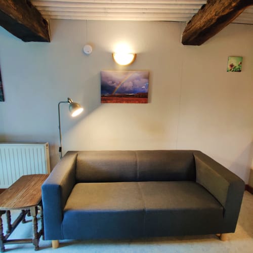 Gezellig zithoekje in de studio van La Ferme De Werpin met donker sofa, houten balken en regenbooglandschapskunstwerk