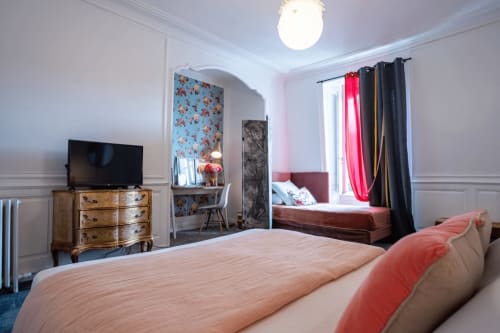 Elegant hotel room at Henri II in Le Mans featuring a bed with coral pillows, wooden dresser with TV, and vintage-style desk