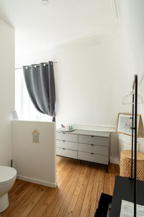 Chambre moderne de l'hôtel Henri II au Mans avec commode blanche, parquets, rideaux gris et entrée salle de bain