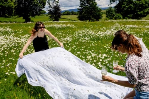Two women holding a white tablecloth in a meadow of white flowers with mountains and trees at Château de Vaux Morvan near