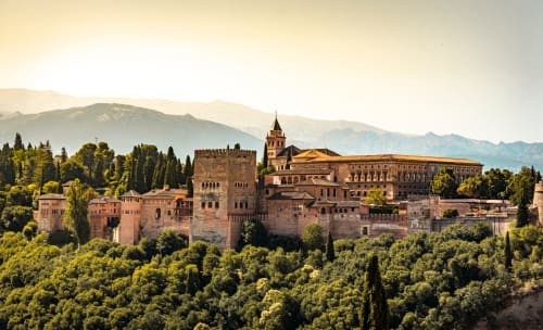 Vista panorámica de la Alhambra de Granada con la Sierra Nevada al fondo, rodeada de vegetación y bañada por la luz dorada.