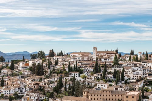 Vista panorámica del Albaicín en Granada, con casas blancas, tejados de terracota, cipreses y montañas al fondo bajo un cielo azul.