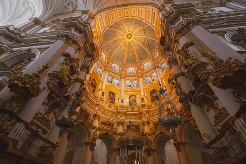 Interior de la Catedral de Granada con su cúpula iluminada, vidrieras, columnas blancas y detalles dorados en estilo renacentista.