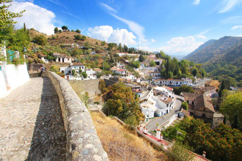 Vista panorámica del Sacromonte en Granada, con casas cueva blancas, colinas verdes y un cielo azul con nubes.