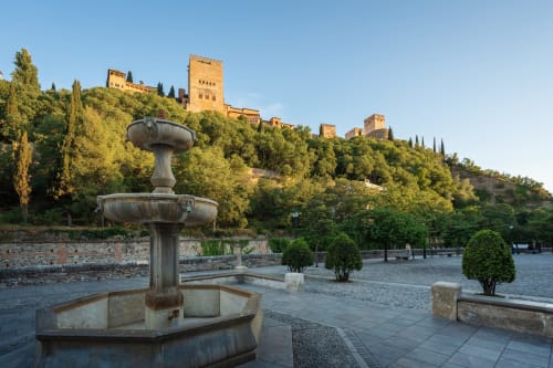 Fuente en Plaza Nueva de Granada con vistas a la Alhambra, rodeada de árboles y con el cielo despejado al atardecer.