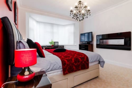Modern king bedroom at Brooklyn House with red accents, flat-screen TV, chandelier, and garden views in Southgate, London.