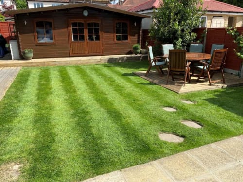 Brooklyn House backyard garden with striped lawn, wooden cabin, patio furniture, and decking in Southgate, London
