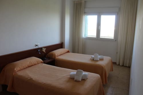 Habitación doble en Apartamentos Los Gemelos en Foz con colchas naranjas, toallas blancas y grandes ventanas con vistas al