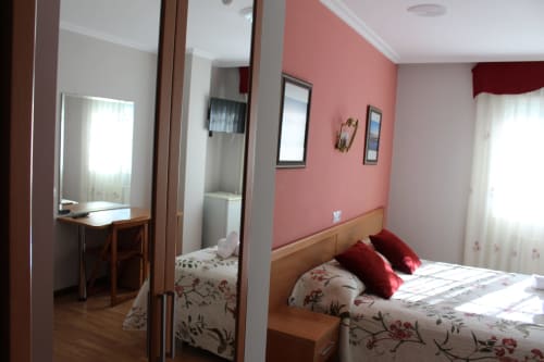 Habitación del Hotel Rego Foz con paredes rosas, cama de madera, almohadas rojas, espejo y ventanas luminosas en Foz, Lugo