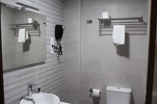 Baño moderno del Hotel Rego Foz con paredes de azulejo gris, ducha de cristal, lavabo blanco, inodoro y toalleros de metal