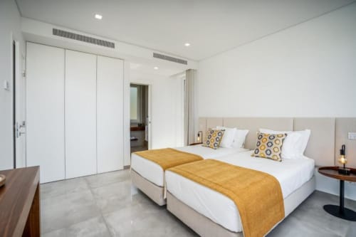 Quarto moderno com duas camas de solteiro, cobertores dourados, cabeceira bege, guarda-roupa branco e decoração neutra na villa Casa Da Colina