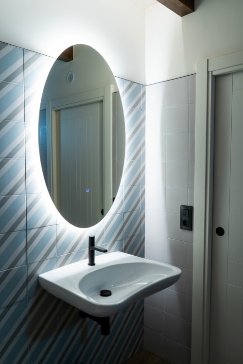 Baño moderno con espejo ovalado iluminado, lavamanos blanco y azulejos a rayas azules en Casa Rural San Andrés de Teixido