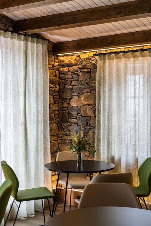 Comedor moderno en Casa Rural San Andres de Teixido con mesa redonda negra, sillas verdes, pared de piedra y cortinas translúcidas