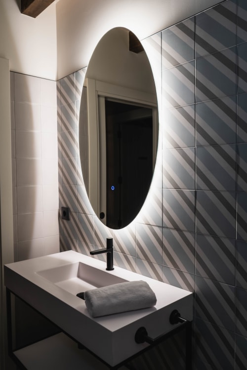 Baño moderno con espejo ovalado iluminado, pared con azulejos rayados, lavabo blanco y grifo negro en Casa Rural San Andres de