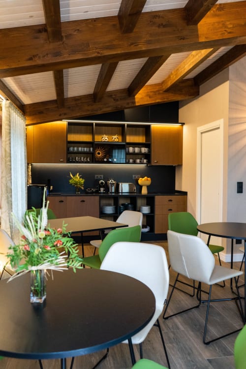 Comedor moderno con mesas redondas negras, sillas verdes y blancas, vigas de madera vista y cocina equipada en Casa Rural