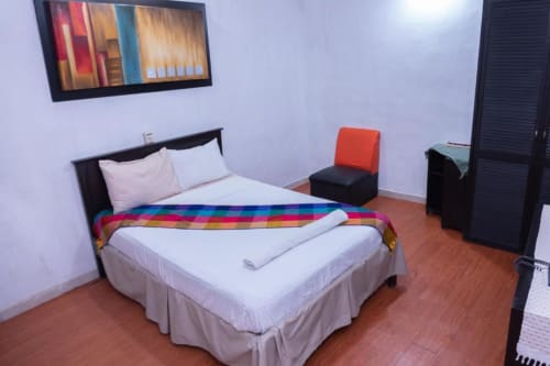 Dormitorio del Hotel Makarios en Tuxtla Gutiérrez con cama, manta colorida, sábanas blancas y silla acentuada naranja